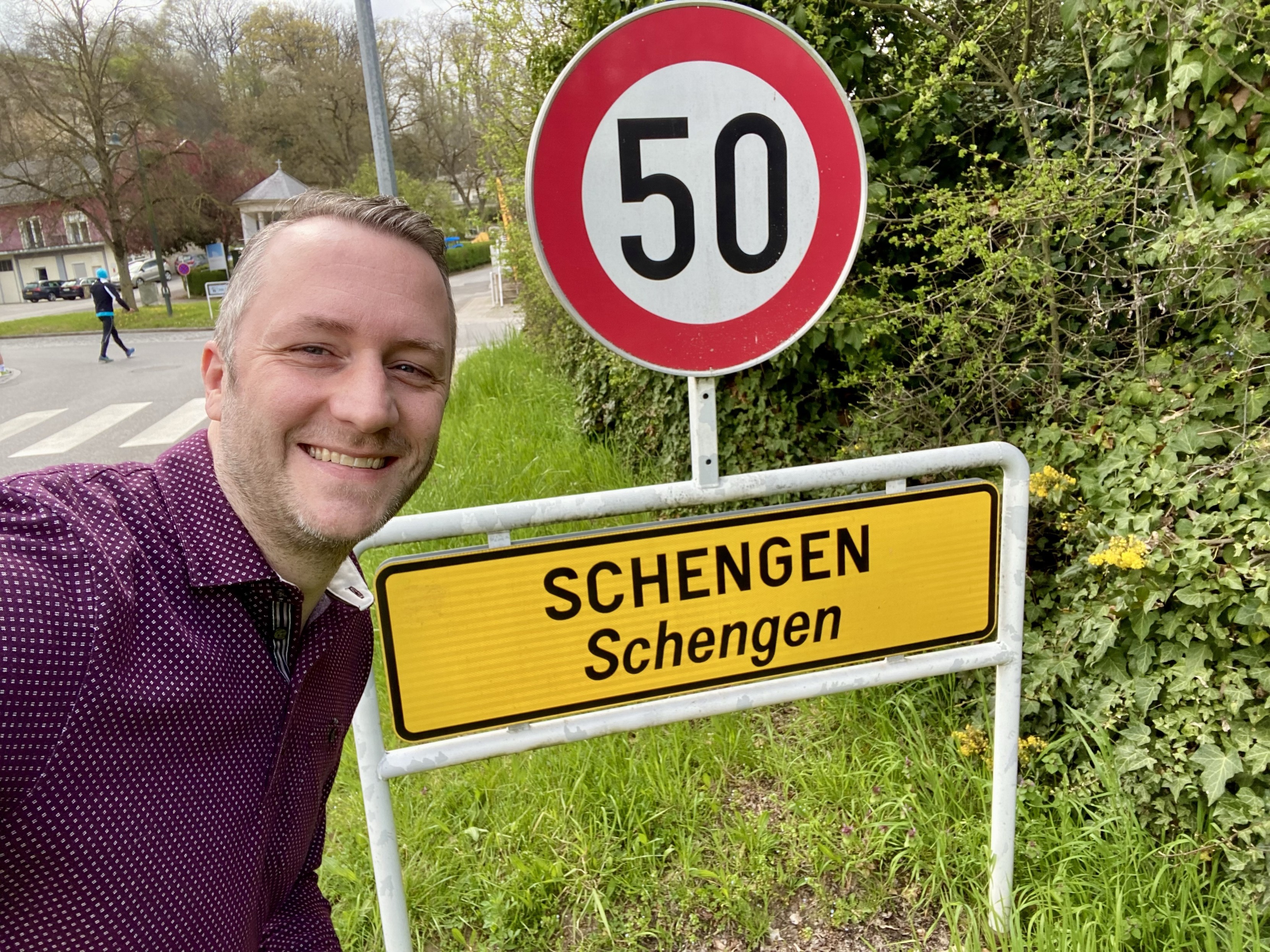 A selfie, I’m wearing a purple/violet top and smile into the camera. I’m standing next to a sign saying we’re about to enter the Luxembourg town of Schengen, an important place in EU history. Above the sign is a another sign indicating a speed limit of 50 km/h. In the background: shrubs, a street, a gazebo and one person crossing the street.