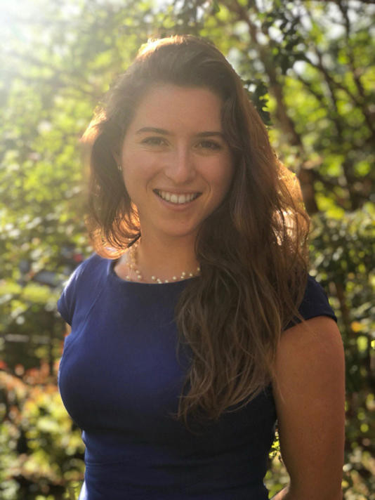 A white woman with long brown hair and a blue dresses looking at the camera while smiling