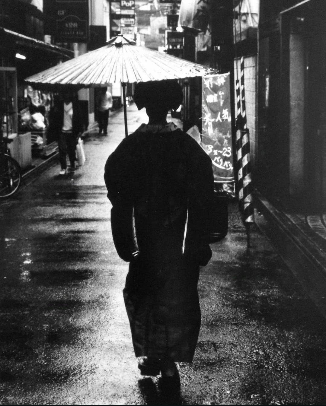 Photograph. A black and white photo of a walking geisha photographed from behind. The young woman is walking with a Japanese umbrella through a small shopping street in the rain. She is wearing a traditional kimono, which can be recognized as a strange silhouette through the dark photograph. Her shoulders are strongly emphasized and form a triangle with the wide sleeves. The waist is narrow and strangely long, it looks like a rectangle. The kimono ends at the legs as a bulbous skirt, which is somewhat irritating. A very nice street photo with a special eye for details.
Info: Fusayoshi Kai (born 1949 in Oita) is a writer and, above all, a photographer who loves and photographs Kyoto. He is the owner of the legendary café "Honyarado", which was destroyed in a fire in 2015, and the "Bar Hachimonjiya", where various cultural figures still meet every evening. He has published over 40 photo books.