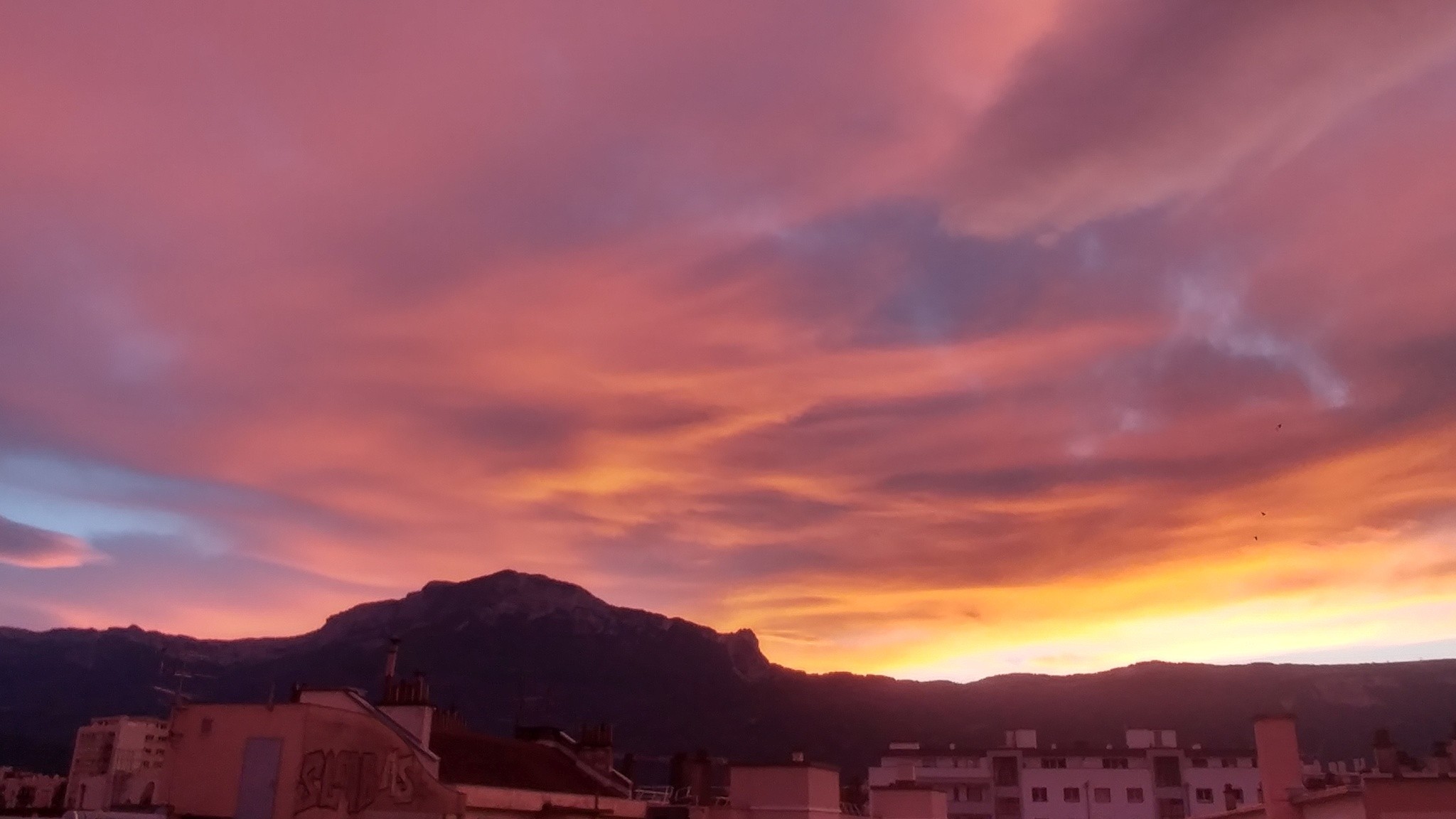 Ciel orange au-dessus du profil d'une montagne qui fait une bosse au milieu mais prend tout l'horizon. Des toits d'immeubles au premier plan, dans une lumière orangée. 