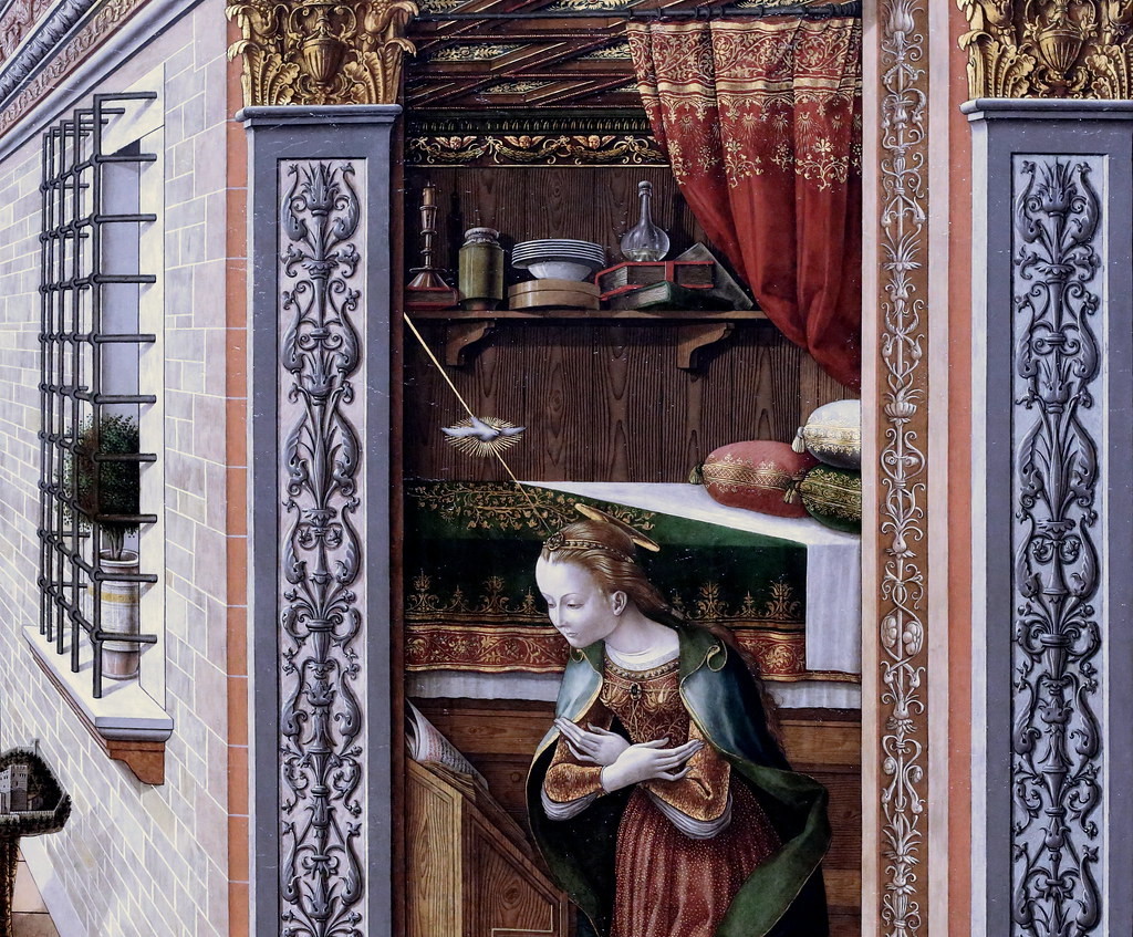 Détail de l'Annonciation de Carlo Crivelli, 1486. Le détail montre la Vierge, agenouillée, les mains croisées sur sa poitrine. En arrière-plan, on voit sa chambre meublée d'un lit et d'une étagère chargée d'objets. La porte de sa chambre est encadrée de pilastres décorées de motifs ornementaux caractéristiques de la période. Sa fenêtre, fermée par une grille, est visible à gauche. Enfin, le Saint Esprit sous la forme d'une colombe arrive au-dessus de sa tête, dans un rayon doré.