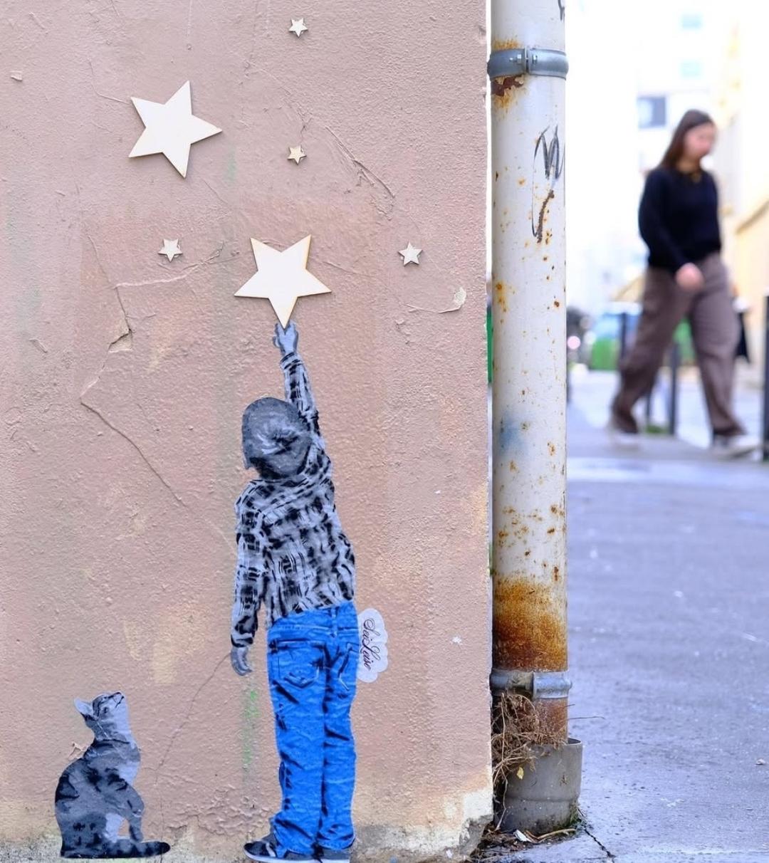 Streetartwall. A beautiful picture and several golden stars have been stuck to the bottom corner of a beige house wall. A small boy in jeans and a blue and white checked shirt is shown from behind, reaching for one of the larger golden stars on the wall.  A black and white cat sits next to him and watches him. (In the photo, a passer-by can be seen somewhat out of focus in the background, which suggests that the mural is no higher than 1.50 m).
INFO: SeiLeise (means in english: "Be Quiet") is a street artist from Cologne. With his colourful paste-ups he has declared war on the grey of the cities and comments on current events; partly playfully, partly with explicit social criticism. Pasteup are duplicated and pasted on walls in several places, often in different countries. 