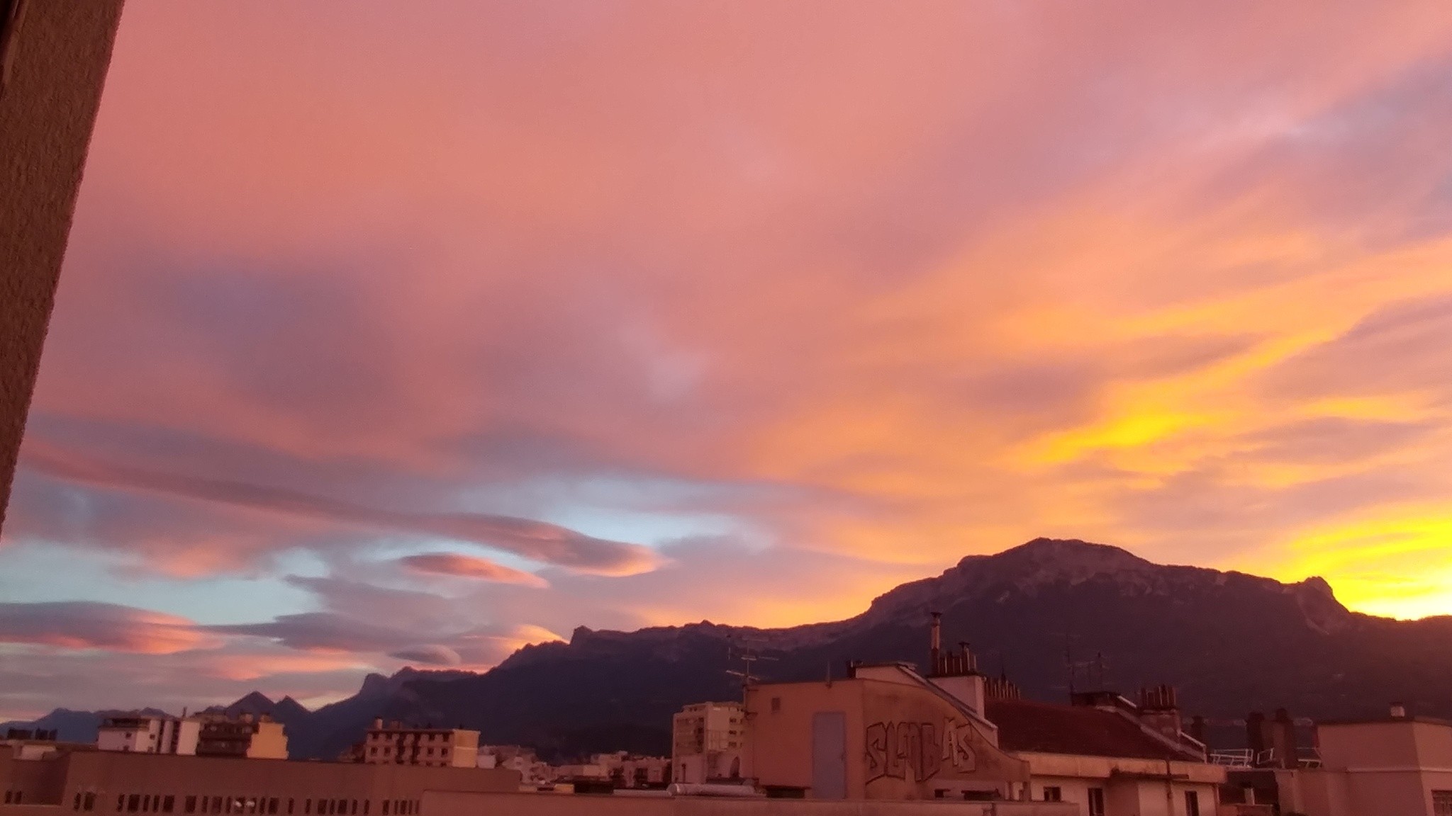 Toits d'immeubles, profil de montagne et nuages en forme de pelotes de coton, orangés. 