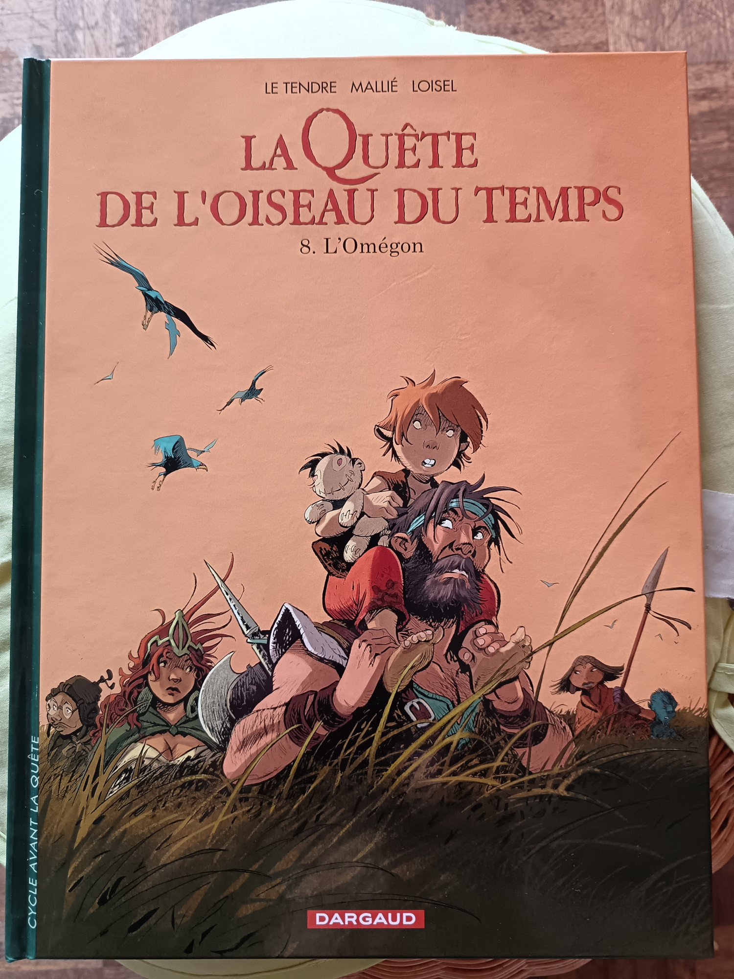 photo : couverture du Tome 8 de la Quête de l'oiseau du temps.
L'illustration montre le chevalier Bragon, marchant dans de hautes herbes, portant sur ces épaule un petit garçon qui tient un doudou dans ses bras. À l'arrière de Bragon progresseent également dans les hautes herbes : Bulrog, Krill, Mara et Bodias.