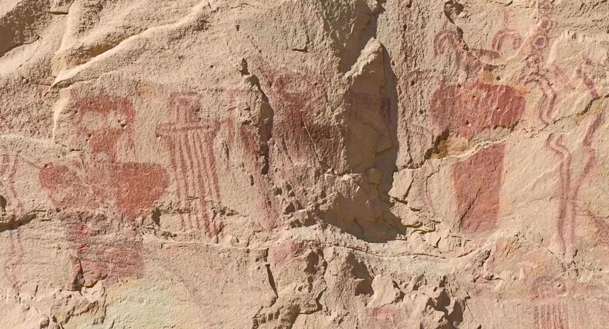 A color landscape photo of a beige / tan sandstone cliff face showing ancient images painted in a brick red ocher. There are five prominent images. On the far left is an anthropomorphic figure of the upper torso and head. There are no arms. There are very large eye holes in the triangular shaped head. The head appears to have ear rings. Moving right is a smaller figure that is similar, less detailed and composed of vertical lines instead of a solid color. In the middle, on a very rough part of the wall, is another figure similar to the one of the left. Because the sandstone is so rough, it is hard to see. Moving further right is another figure, a bit smaller than the one of the far left. It is very similar in design as the one on the far left although it has twin curled horns rather than ear rings. And on the far right are three wavy lines forming a sort of funnel shape. Two parallel lines on the left and one on the right. Above the two parallel lines on the left is a series of four small circles with large dots in the middle.